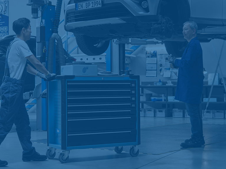 Mechanic rolls workshop trolley to a jacked-up Asian vehicle, where a second mechanic is waiting for a spare part