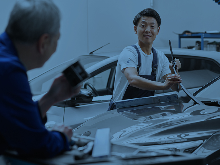 Two mechanics on a closed bonnet, one of whom is fitting a wiper blade